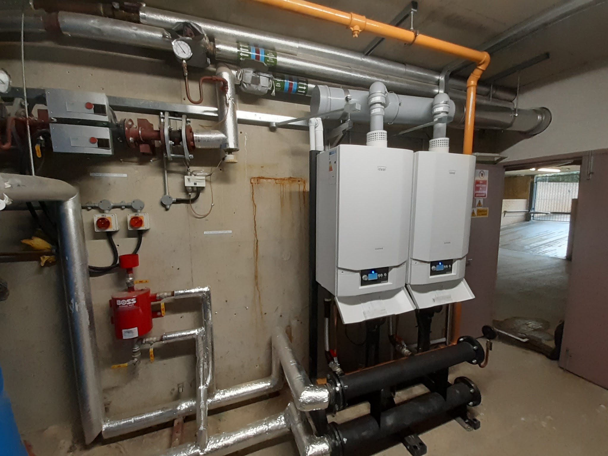 Two wall-hung boilers in a plant room with gas pipework, a header and isolation valves